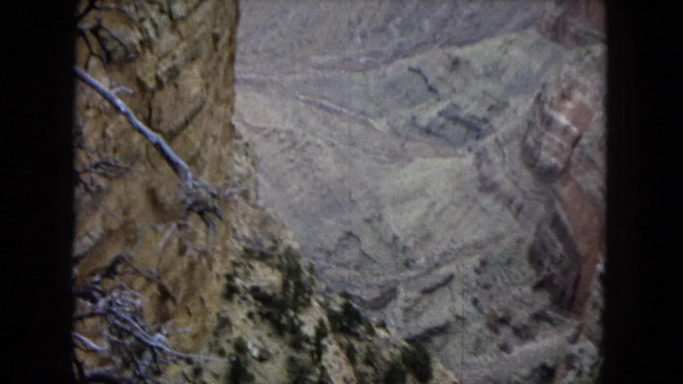 1967 Arizona 8mm frame showing a canyon overlook view with layered rock, pale eroded slopes, dry brush, and dark gate edges from the film scan.