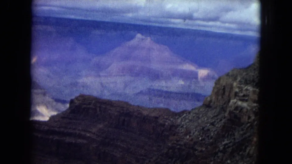 1967 Arizona Grand Canyon aerial frame with distant plateau formations, a shadowed gorge, rocky slopes, and blue-purple color shift from the 8mm film scan.