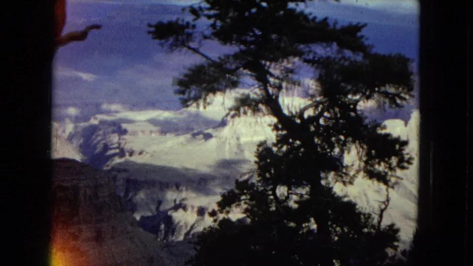 1967 Arizona home-movie footage frames a snowy mountain and canyon view through a dark pine silhouette, with blue-toned sky and visible film wear.