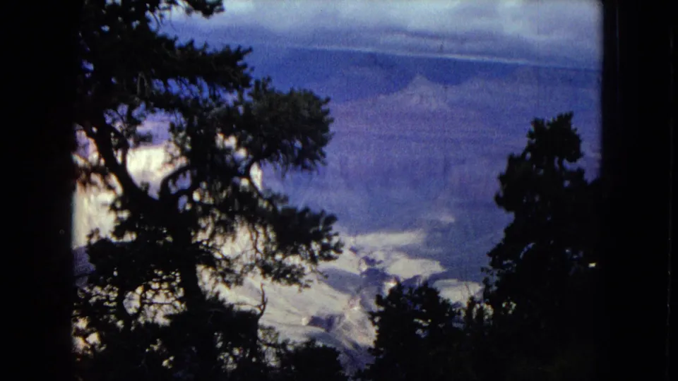 The final 8mm frame places dark pine and tree silhouettes in the foreground against a distant snow-dusted mountain and canyon view under a blue-gray sky.