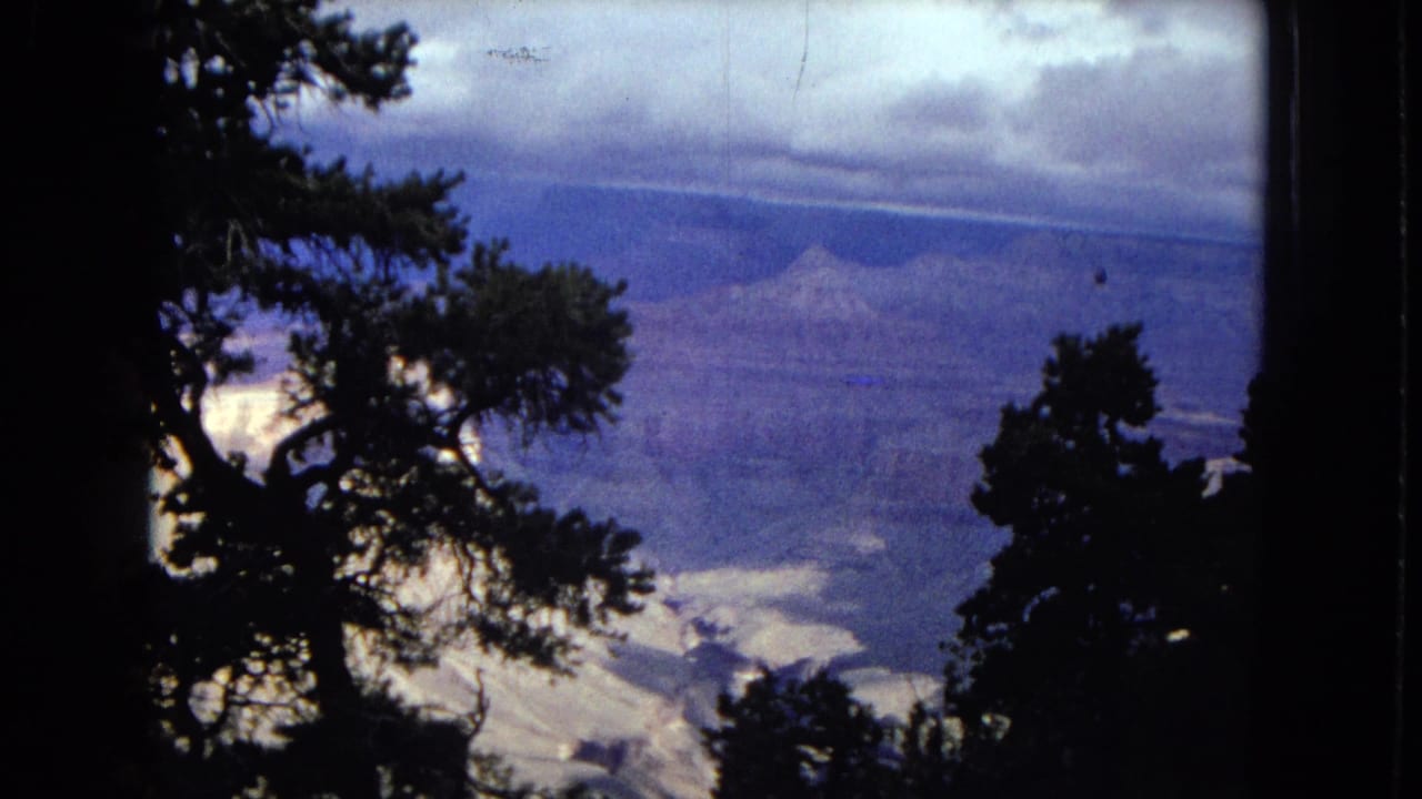 1967 8mm home-movie view of the Grand Canyon in Arizona, seen through dark pine silhouettes with layered canyon walls beneath a cloudy blue sky.