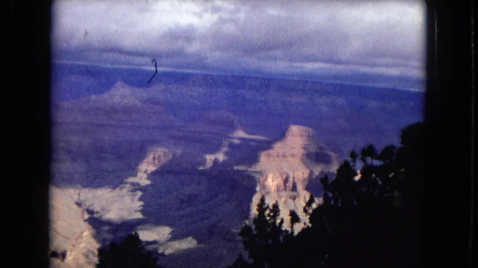 1967 8mm home-movie view across the Grand Canyon, with pale rock buttes, blue shadowed canyon layers, and dark foreground trees.