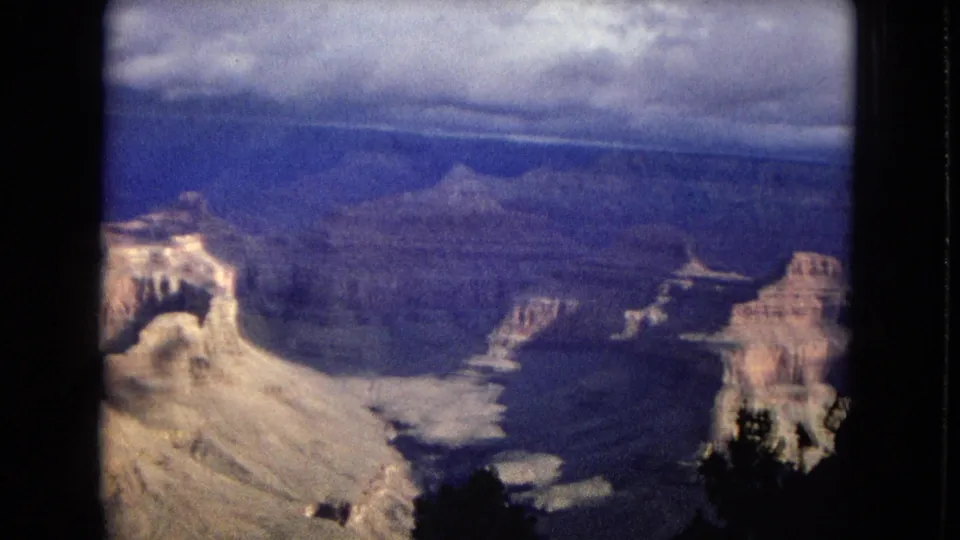 1967 8mm home-movie frame looking over the Grand Canyon in Arizona, showing layered cliffs, a distant mesa, clouded sky, and dark tree silhouettes at the edge.