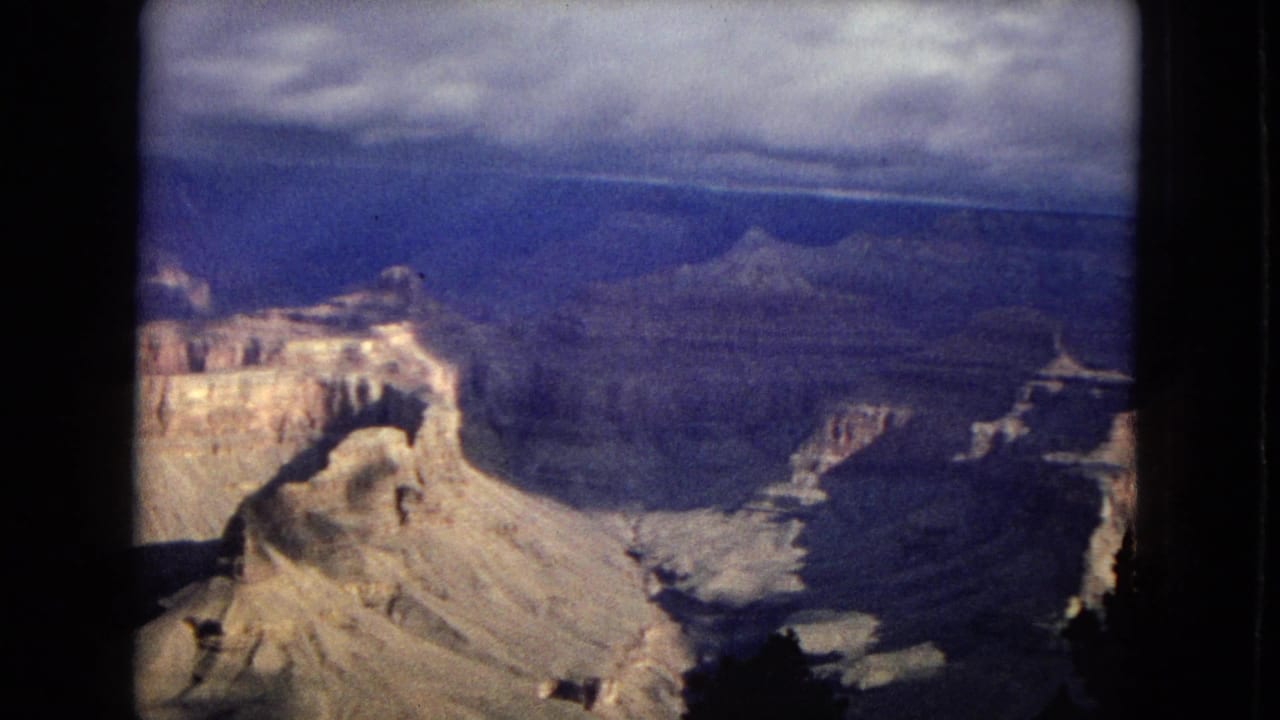 1967 Arizona 8mm frame showing layered Grand Canyon formations, distant blue ridges, and a low cloudy sky from a visitor overlook perspective.