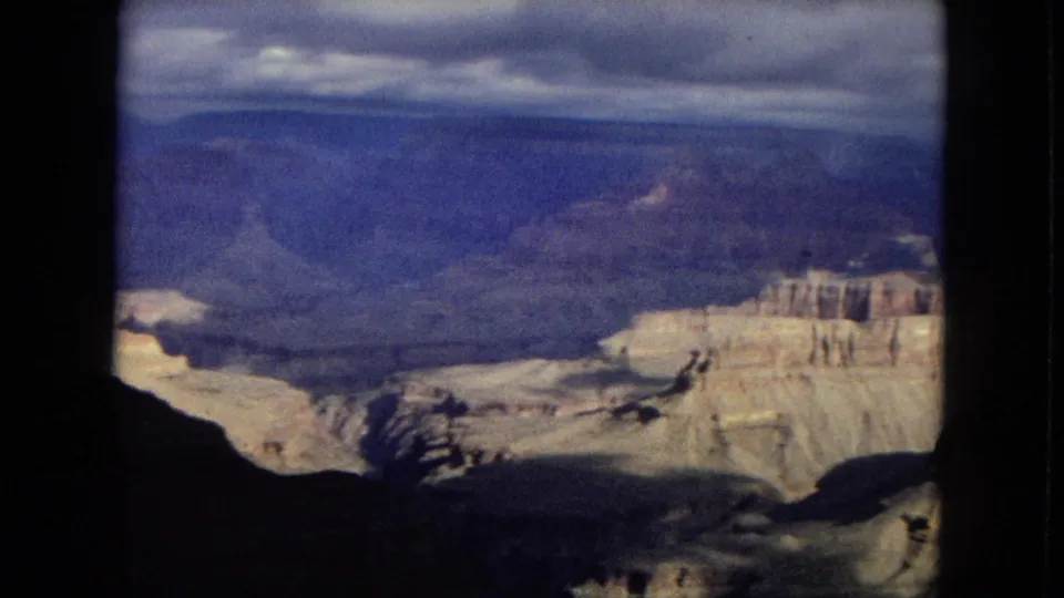 1967 Arizona 8mm frame capturing a wider Grand Canyon panorama with a bright rock ledge, deep shadowed foreground, distant ridgelines, and overcast sky.