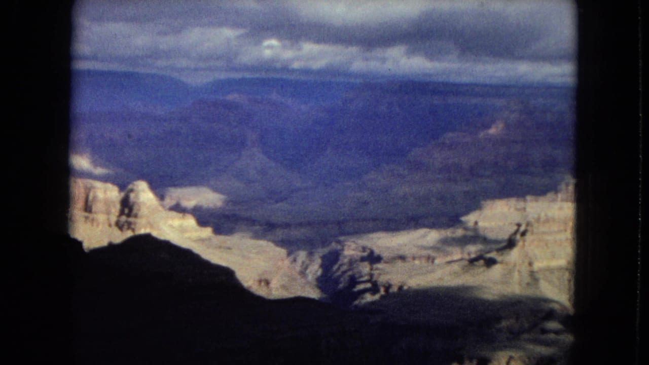 1967 8mm home-movie view of the Grand Canyon with pale layered cliffs, blue distant ridges, heavy clouds, and dark foreground canyon shapes.