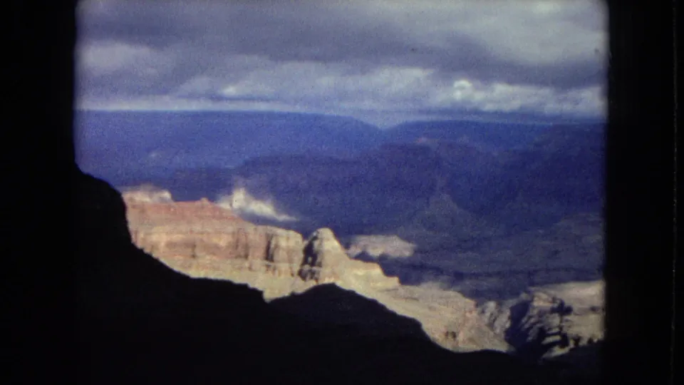 Visitor-shot Grand Canyon panorama from 1967, showing a dark foreground cliff, sunlit layered rock, distant canyon walls, and low clouds.