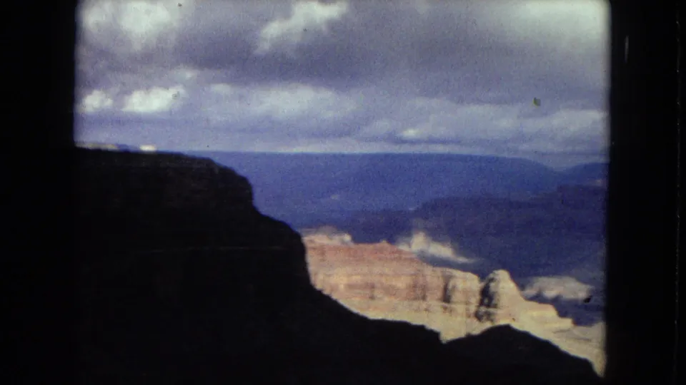1967 Grand Canyon home-movie frame with a dark overlook edge in front of pale cliffs, distant blue canyon layers, and a moody cloud cover.