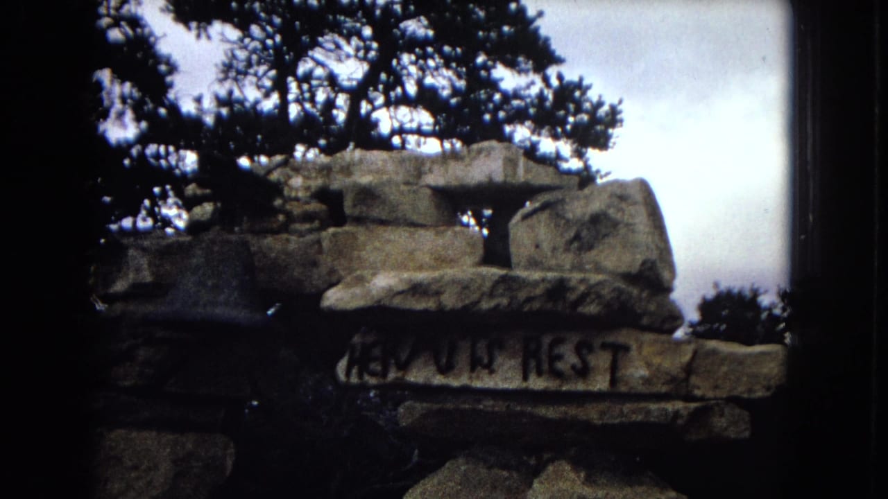 1967 Arizona 8mm frame showing a rough stone structure with painted text that appears to read "HEAVEN REST" under dark evergreen branches and bright sky.