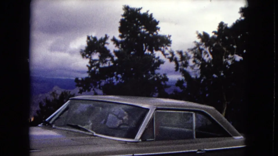 Closer 8mm frame of a light-colored older car at an Arizona scenic overlook, with branches and hazy canyon terrain in the background.