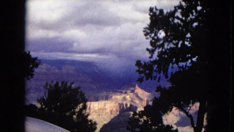 1967 Arizona home-movie frame showing a Grand Canyon vista through silhouetted trees, with layered rock formations beneath an overcast sky.