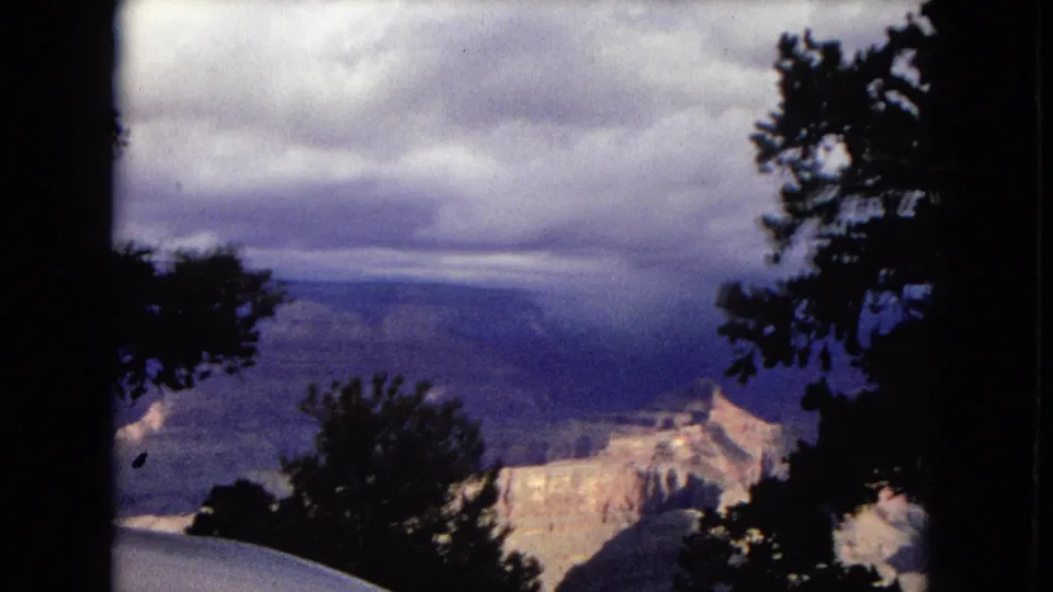 Grand Canyon overlook from 1967 Arizona footage, with distant ridges and pale rock formations framed by dark foreground trees.