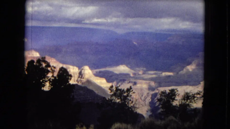 1967 8mm frame of a broad Grand Canyon view with layered rock, distant plateau, foreground shrubs, and an overcast sky.
