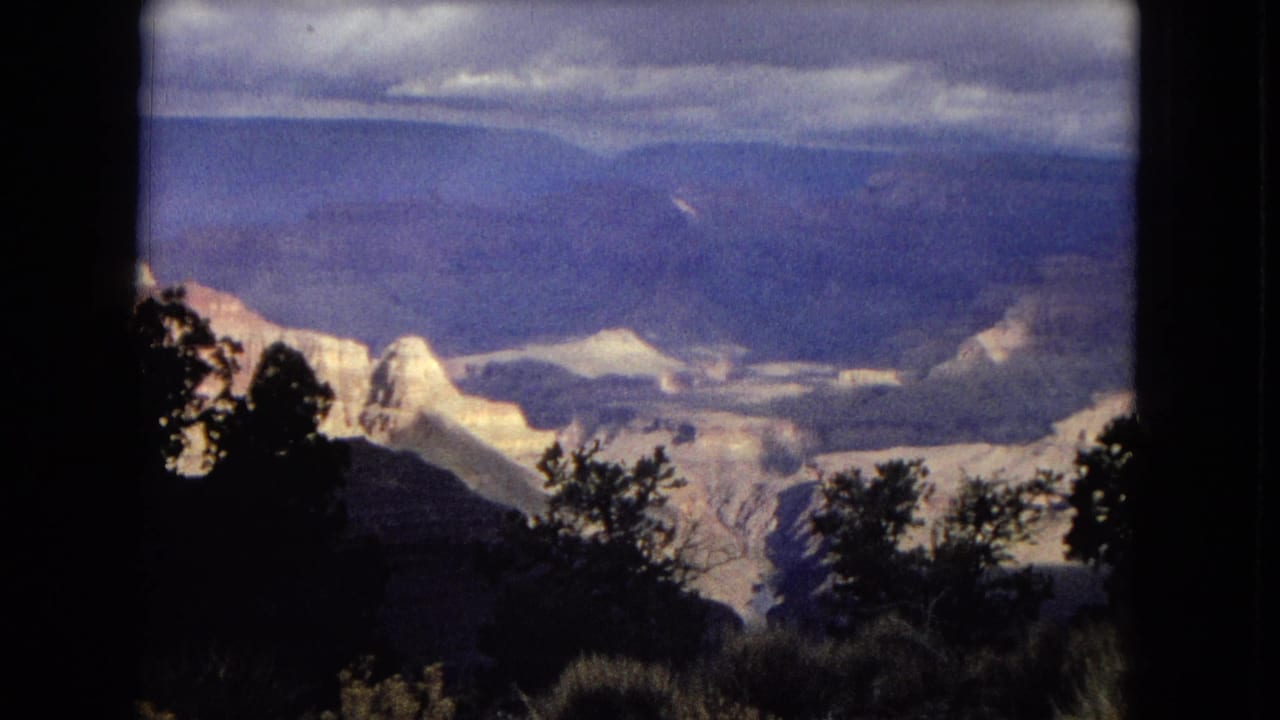 1967 8mm footage frame showing a Grand Canyon overlook in Arizona, with silhouetted foreground trees, pale rock formations, distant canyon ridges, and a cloudy sky.