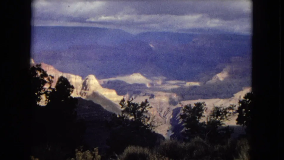 1967 8mm footage frame showing a Grand Canyon overlook in Arizona, with silhouetted foreground trees, pale rock formations, distant canyon ridges, and a cloudy sky.
