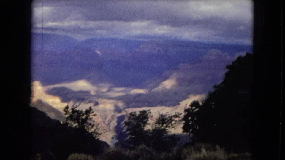 1967 8mm Arizona canyon frame with shrubs and a dark slope in the foreground, pale Grand Canyon formations across the middle distance, and blue ridges below an overcast sky.