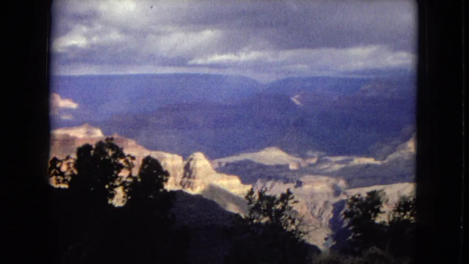 1967 8mm frame from a Grand Canyon overlook, showing dark foreground trees, layered sunlit canyon rock, a broad blue ridge line, and low clouds across the sky.