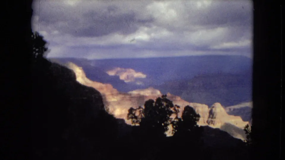 1967 8mm Arizona frame with a dark cliff face and silhouetted trees framing bright Grand Canyon rock formations and distant shadowed ridges under thick clouds.