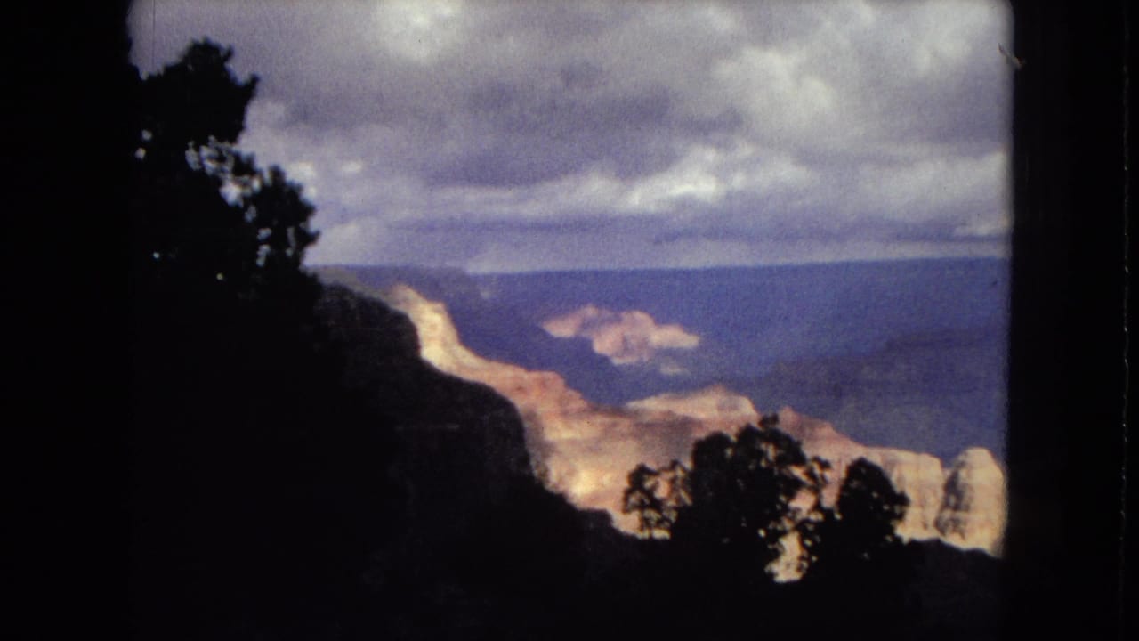 1967 8mm home-movie view of the Grand Canyon, with dark foreground trees framing layered canyon walls beneath an overcast sky.