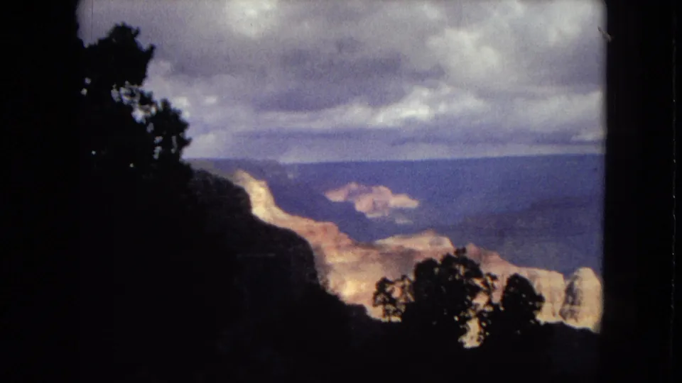 1967 8mm home-movie view of the Grand Canyon, with dark foreground trees framing layered canyon walls beneath an overcast sky.