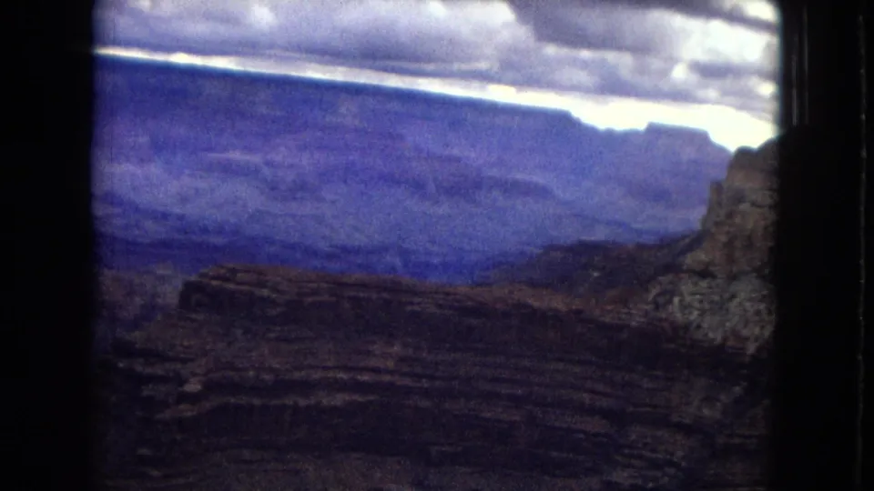 Wide 1967 Grand Canyon home-movie view across dark layered valley walls toward distant mesas beneath a moody cloud band.