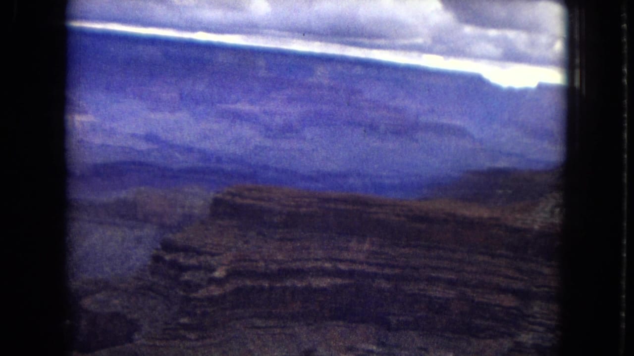 1967 8mm home-movie frame showing an aerial view over Grand Canyon-like layered rock formations beneath a cloudy sky with visible film borders.