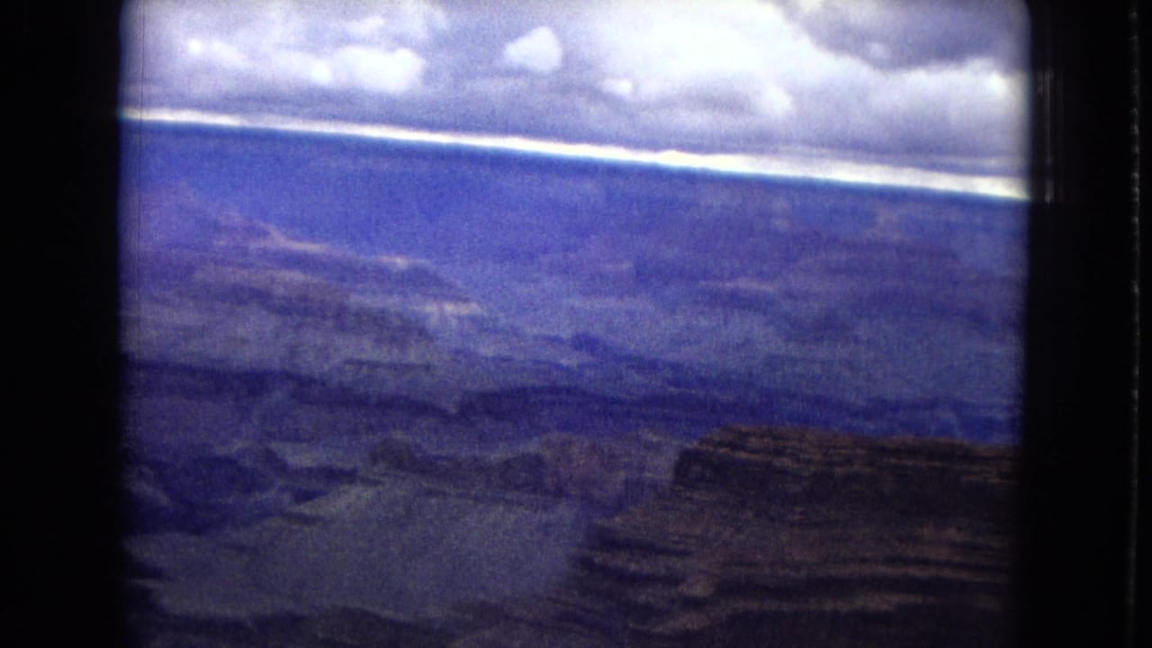 Wide aerial canyon view with mesas, ravines, and a bright cloud band at the horizon.