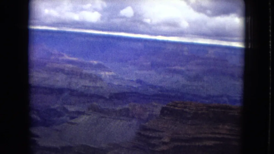 1967 8mm aerial travel frame looking across broad Grand Canyon terrain, with distant ravines, flat-topped rock formations, and an aged blue-purple film cast.