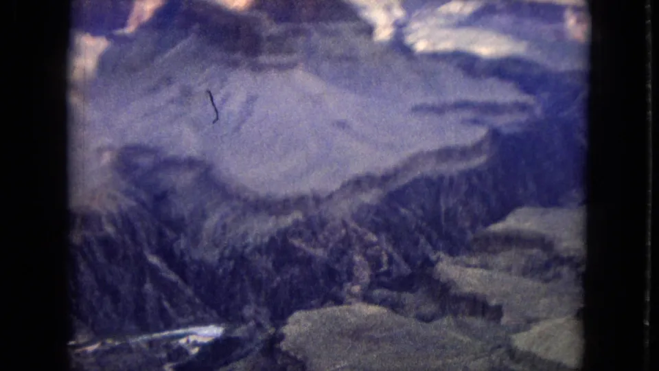 1967 8mm aerial frame looking down into a deep Grand Canyon basin, with ridged rock walls, pale shelves, and visible scratches and grain from the film scan.