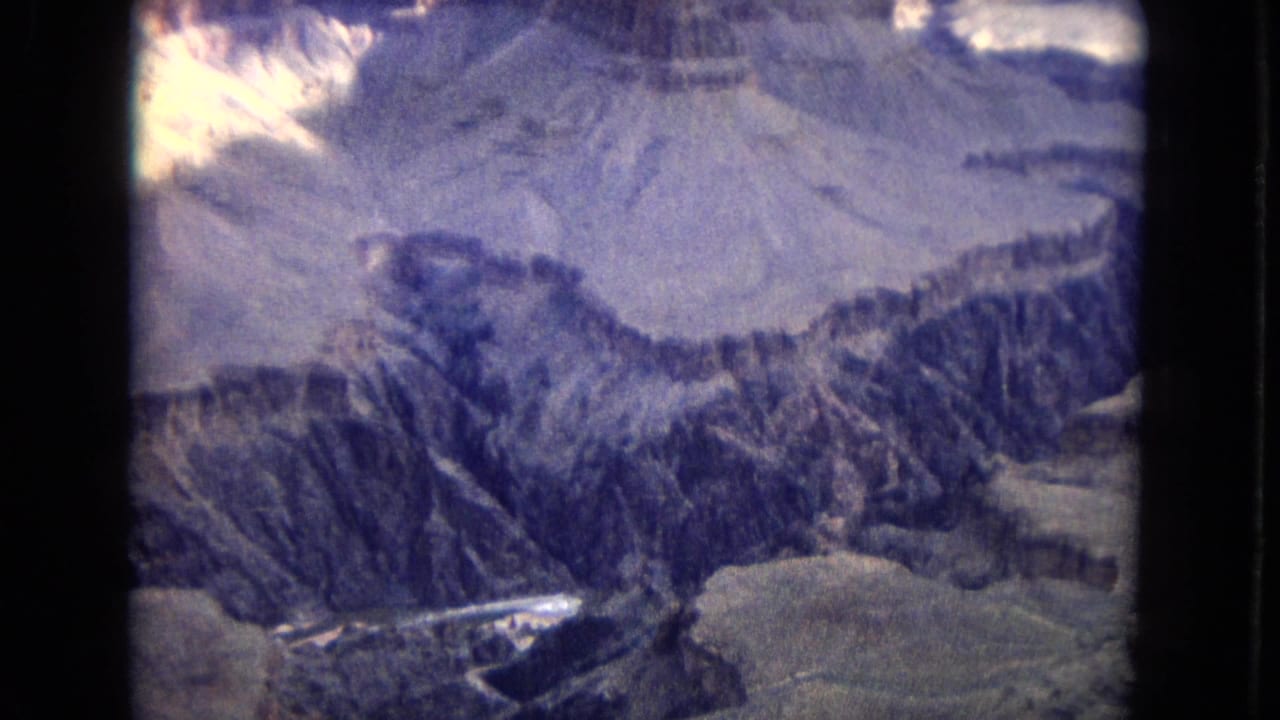 1967 8mm aerial view over the Grand Canyon area, showing layered rock walls, pale mesa surfaces, and a narrow watercourse below with aged color-film grain.