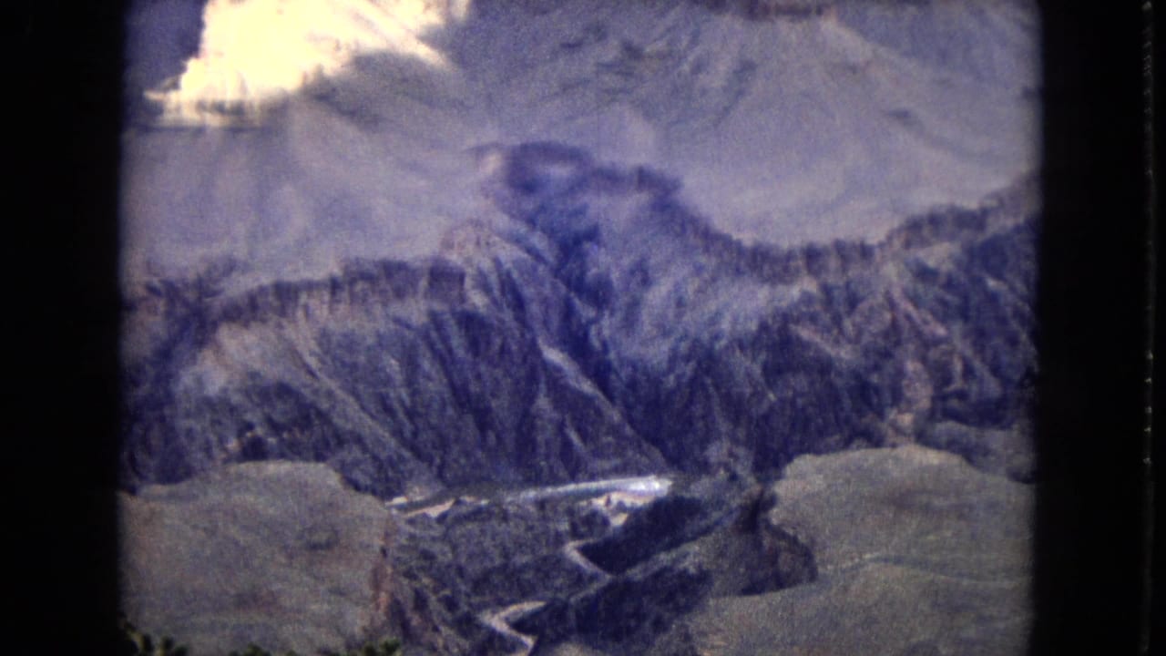 1967 8mm aerial view of Grand Canyon terrain with steep ridges, shadowed chasms, pale upper landforms, and a winding stream channel visible near the bottom edge.