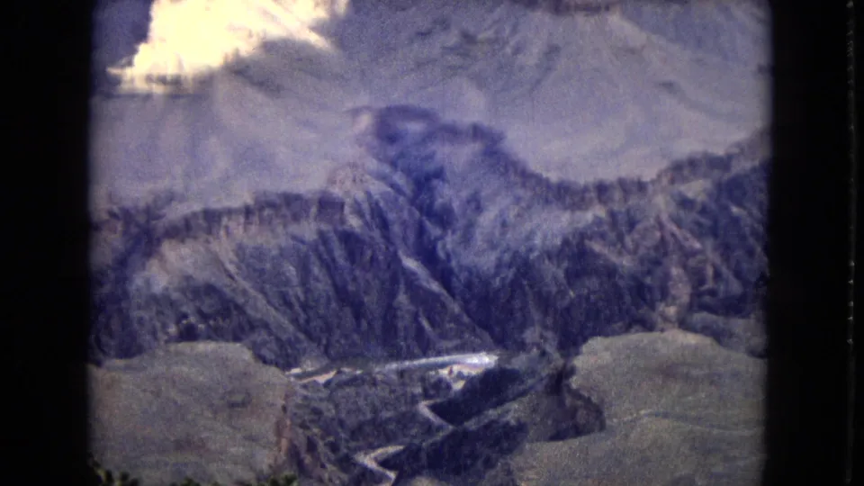 1967 8mm aerial view of Grand Canyon terrain with steep ridges, shadowed chasms, pale upper landforms, and a winding stream channel visible near the bottom edge.