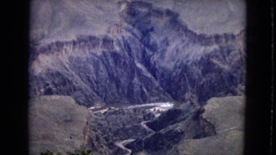 1967 8mm aerial view looking down across rugged Grand Canyon walls, with striated slopes, pale mesa edges, and a bright winding watercourse at the canyon floor.
