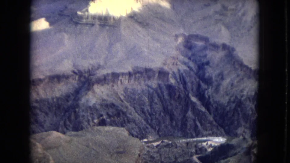 1967 8mm aerial view across Grand Canyon cliffs and eroded slopes, with pale foreground rock, a narrow channel below, and visible dark borders from the scanned film frame.