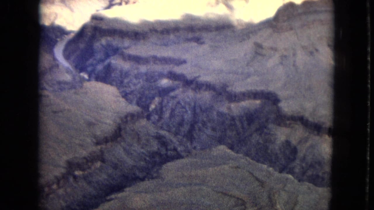 A high view across the Grand Canyon landscape, with pale rock terraces, shadowed canyon walls, and the river visible in the distance on faded 8mm film.