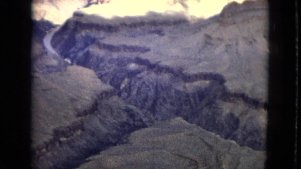 A high view across the Grand Canyon landscape, with pale rock terraces, shadowed canyon walls, and the river visible in the distance on faded 8mm film.