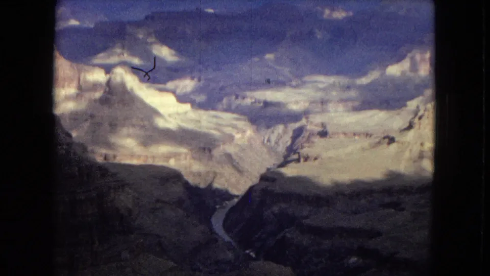 Wide 1967 8mm view across the Grand Canyon under a cloudy sky, with pale rock layers, dark canyon slopes, and the river cutting through the center distance.