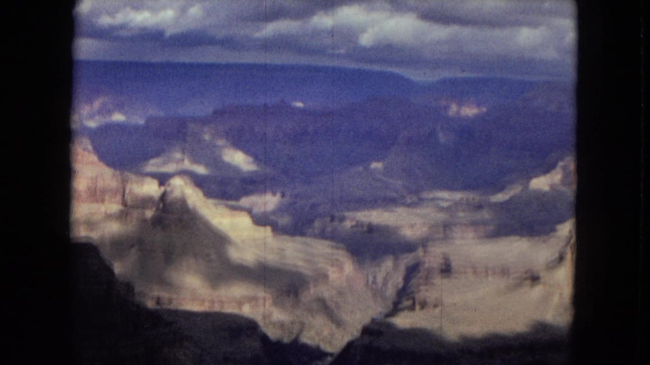 1967 Arizona 8mm frame showing a broad Grand Canyon vista with layered pale rock formations, dark foreground shadow, distant blue plateau, and cloud-filled sky.