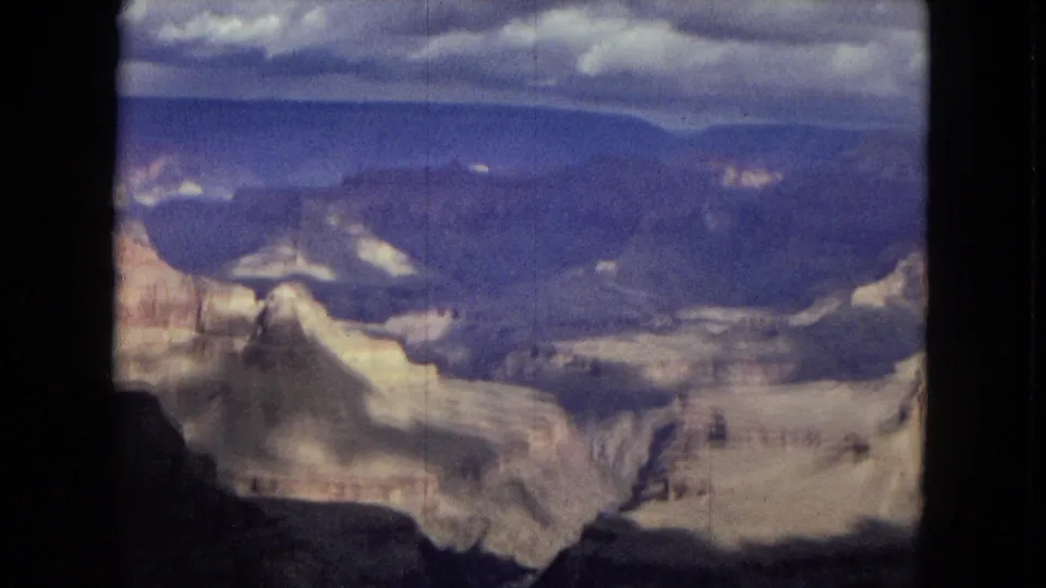 1967 Arizona 8mm frame showing a broad Grand Canyon vista with layered pale rock formations, dark foreground shadow, distant blue plateau, and cloud-filled sky.