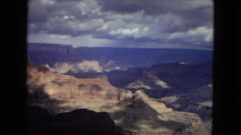 1967 Arizona 8mm frame looking across the Grand Canyon toward sunlit ledges, dark ravines, distant mesas, and a dense cloud bank overhead.