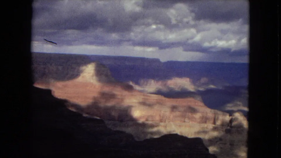 1967 Arizona 8mm frame with a dark canyon rim in the foreground, broad reddish and pale rock formations, distant canyon walls, and a gray-blue cloudy sky.