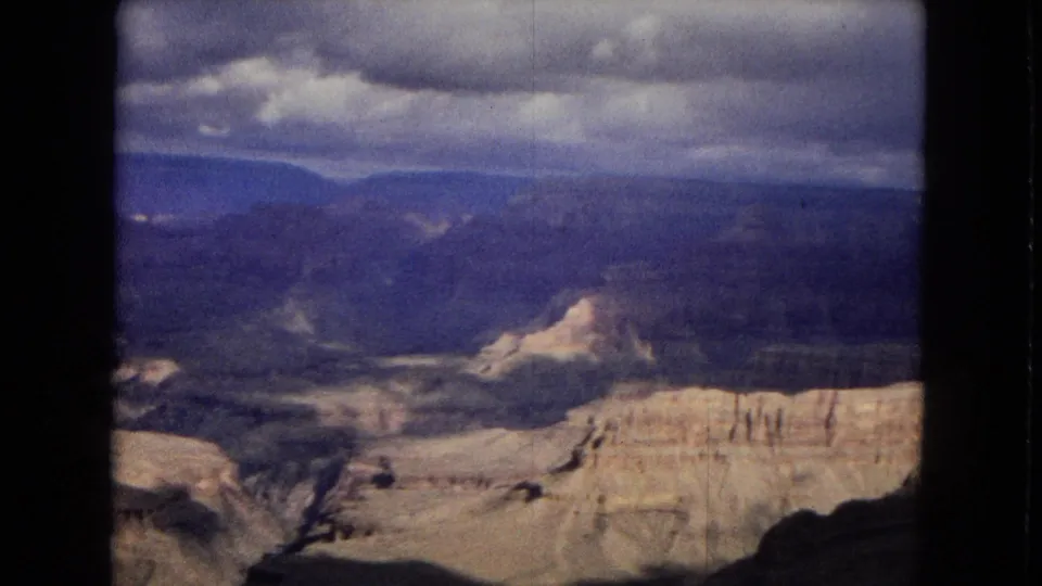 1967 Arizona 8mm frame showing a distant Grand Canyon basin with pale stratified rock, shadowed valleys, blue mesas on the horizon, and heavy clouds above.