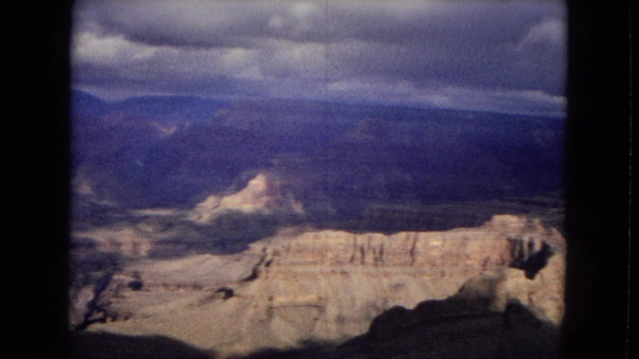 1967 8mm home-movie frame showing sunlit Grand Canyon rock layers beneath a heavy cloudy sky, with dark scan borders visible at the sides.