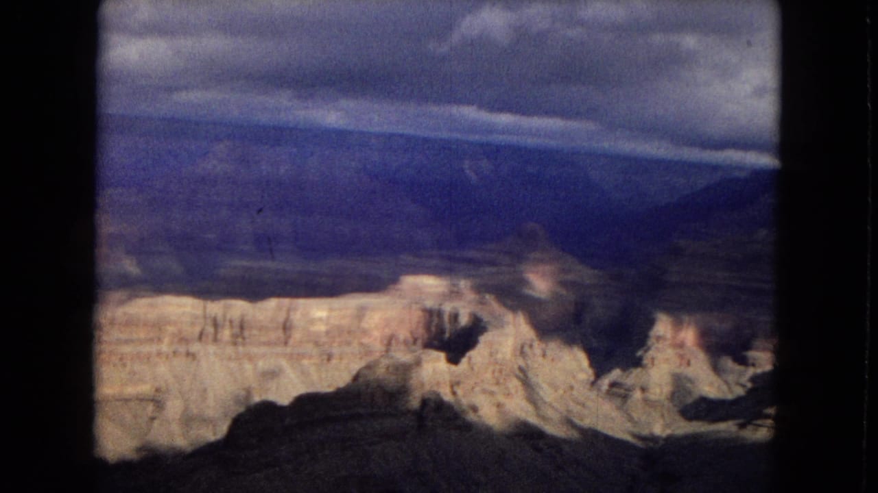 1967 8mm home-movie frame looking across the Grand Canyon, with pale rock terraces, dark shadowed slopes, distant blue mesas, and cloud cover.