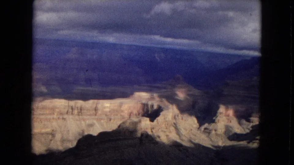 1967 8mm home-movie frame looking across the Grand Canyon, with pale rock terraces, dark shadowed slopes, distant blue mesas, and cloud cover.