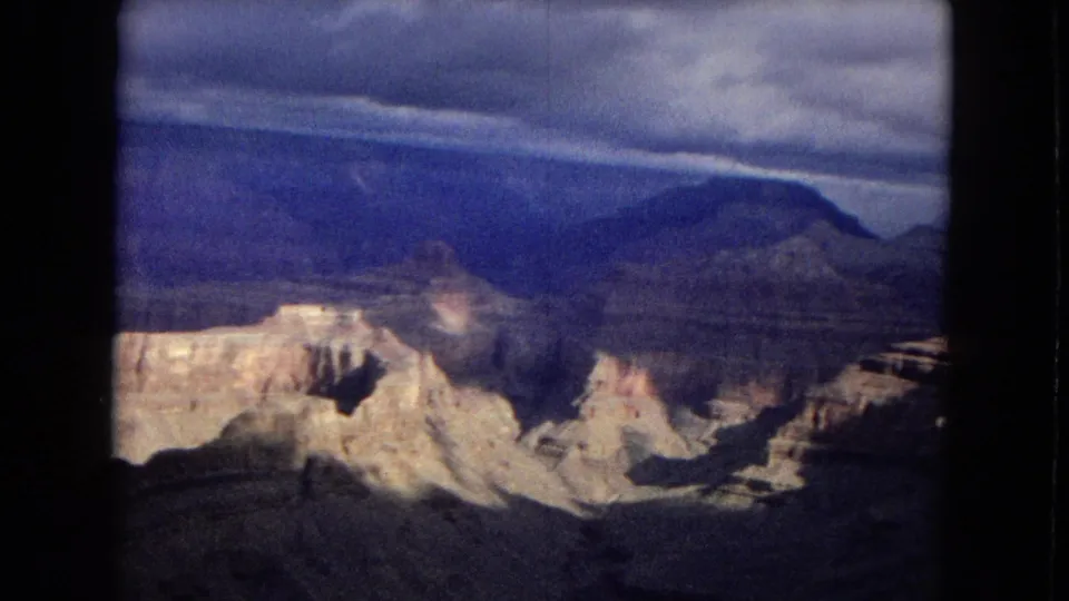 1967 8mm home-movie frame showing sunlit canyon formations in the lower frame, a dark blue mesa beyond, and a textured cloudy sky above.