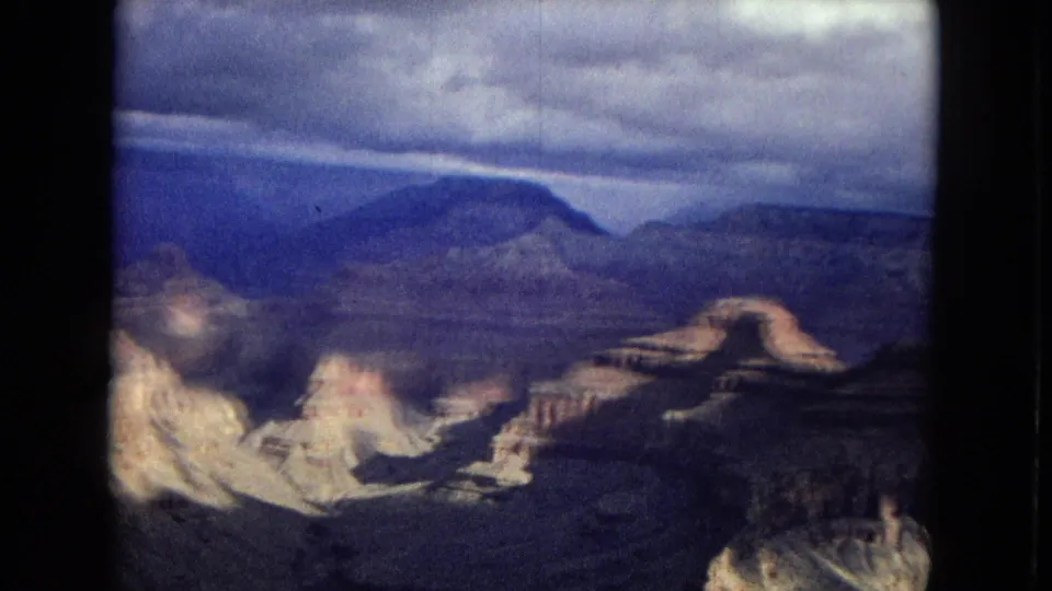 1967 8mm home-movie frame showing Grand Canyon ridges, pale eroded rock formations, distant mesas, and a cloudy sky with visible film softness and grain.