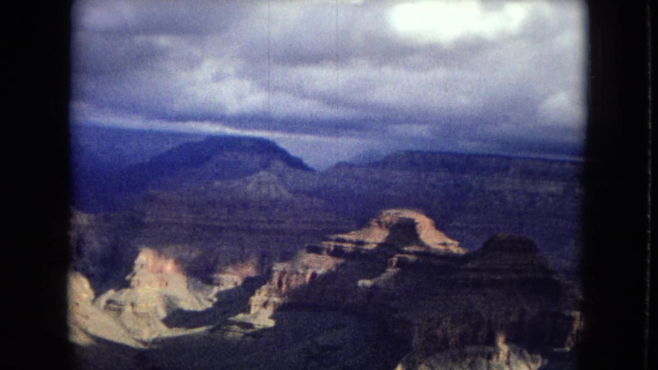 1967 8mm home-movie frame of the Grand Canyon from a high overlook, showing layered canyon formations beneath a dark, overcast sky.