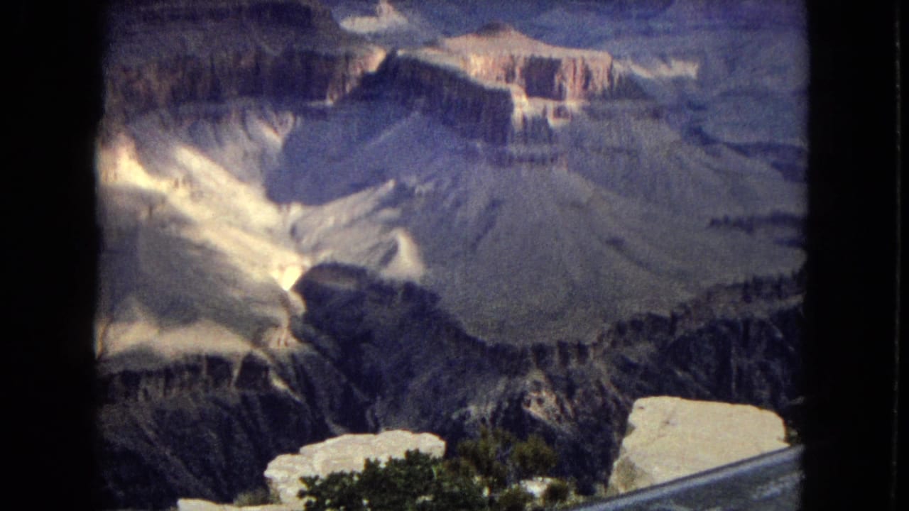 1967 Grand Canyon overlook frame showing shadowed ravines, pale rock layers, storm clouds, and a visible metal guardrail in the foreground.