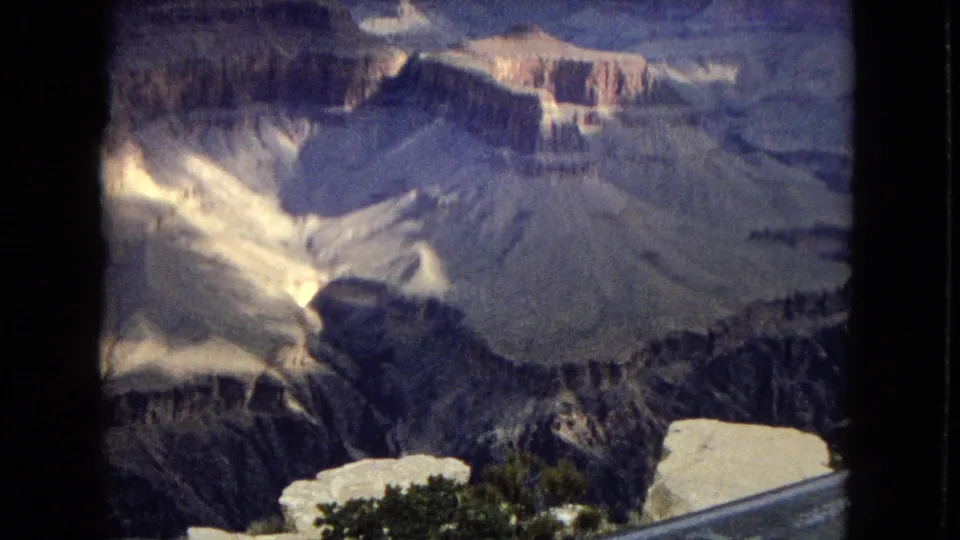 1967 Grand Canyon overlook frame showing shadowed ravines, pale rock layers, storm clouds, and a visible metal guardrail in the foreground.